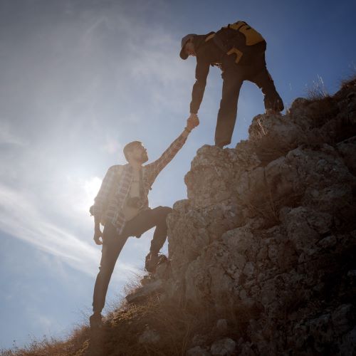 A person reaches down to assist another person in climbing up a large rock